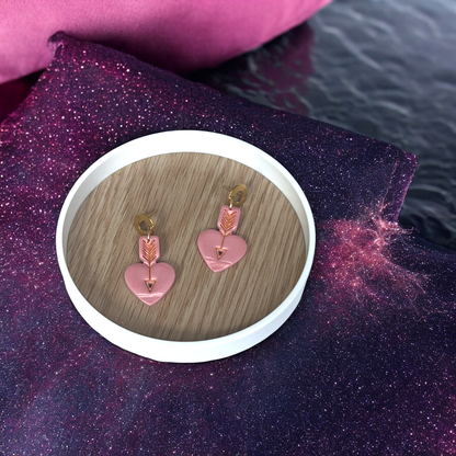 Pink heart-shaped drop earrings with gold arrows and circular studs, placed on a wooden tray against a glittery cosmic fabric backdrop.