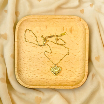 Gold ribbed heart pendant necklace with a shiny finish displayed on a wooden tray against a soft beige backdrop featuring heart patterns.