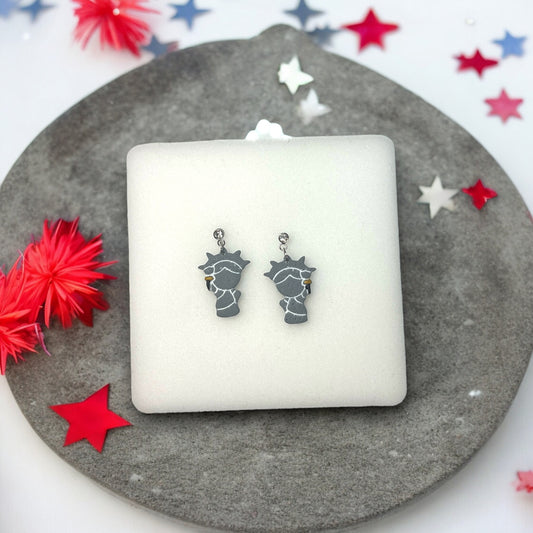 Gray polymer clay Statue of Liberty dangle earrings with rhinestone studs, displayed on a soft white cushion with festive decor.
