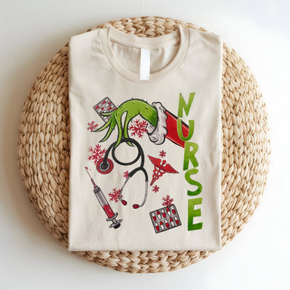 Flat lay of a beige Grinch-themed nurse t-shirt featuring holiday medical graphics like stethoscopes, syringes, and festive decor.
