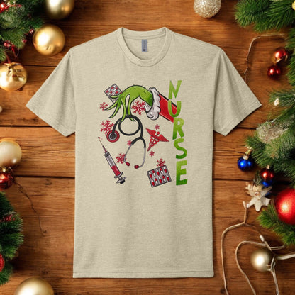 Flat lay of a beige Grinch-themed nurse t-shirt featuring holiday medical graphics like stethoscopes, syringes, and festive decor.