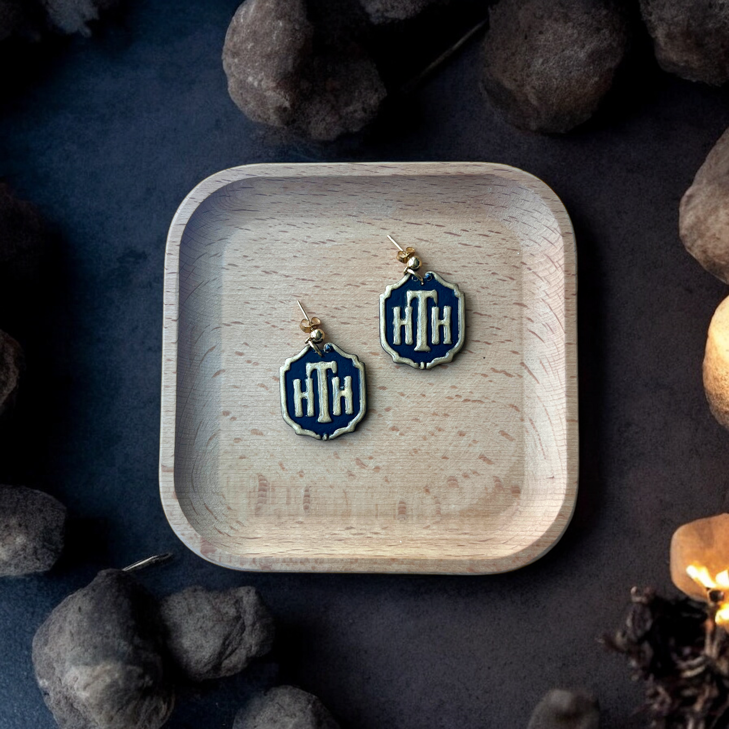 Pair of HTH hotel plaque-inspired earrings with gold accents placed inside a square wooden tray, surrounded by stones and a dim, eerie setting.
