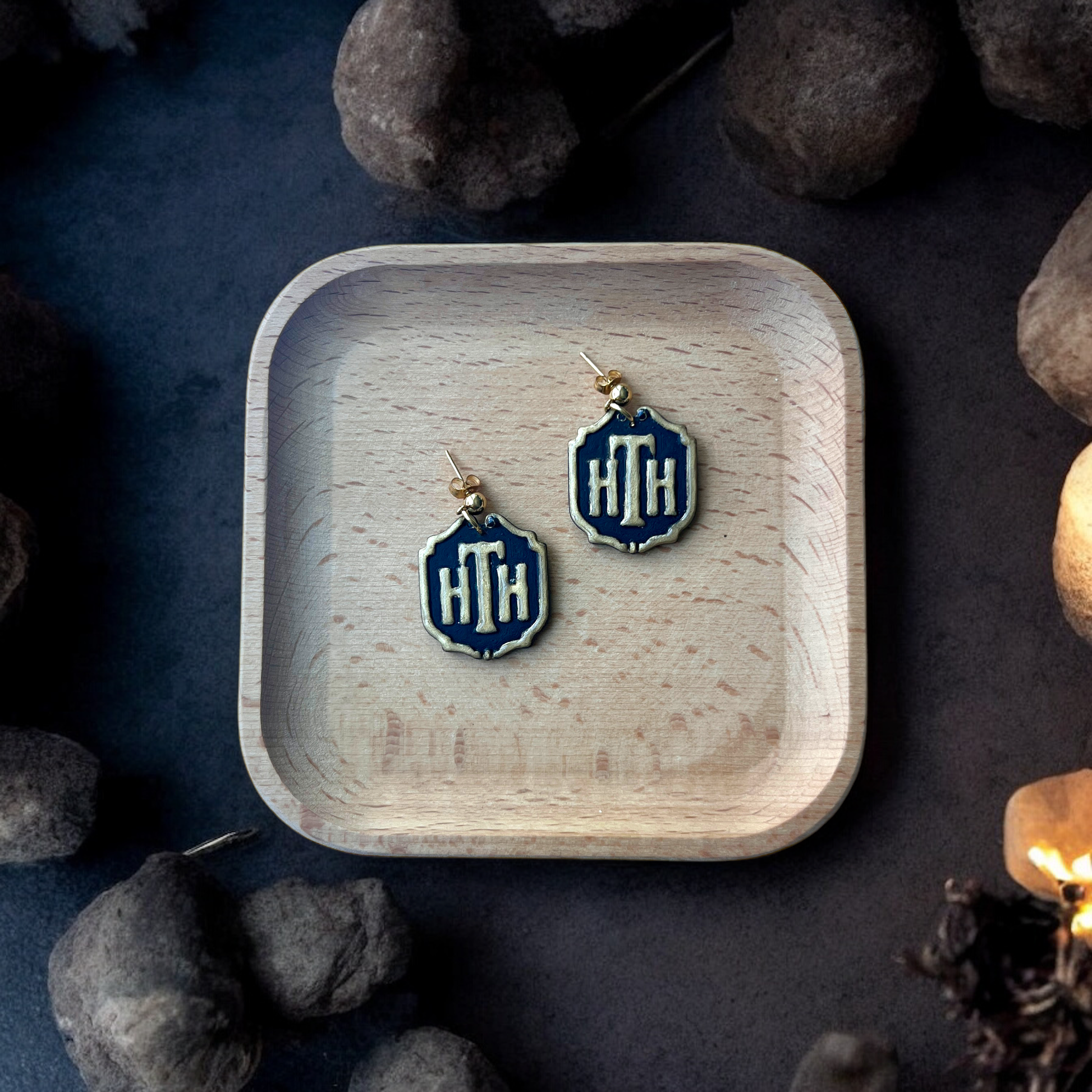 Pair of HTH hotel plaque-inspired earrings with gold accents placed inside a square wooden tray, surrounded by stones and a dim, eerie setting.