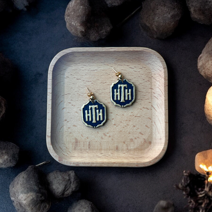 Pair of HTH hotel plaque-inspired earrings with gold accents placed inside a square wooden tray, surrounded by stones and a dim, eerie setting.