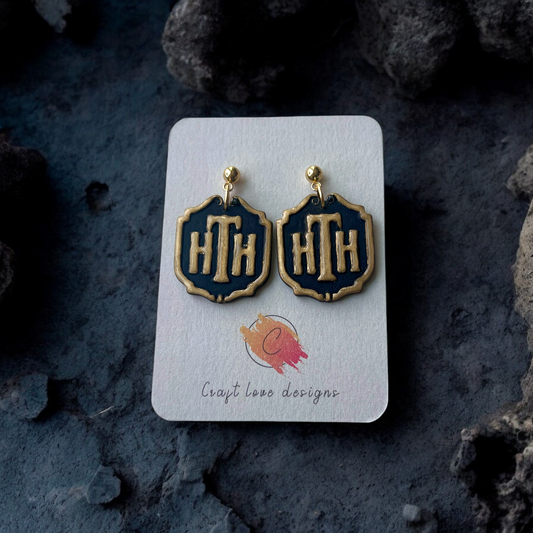 Pair of black and gold HTH hotel plaque-themed earrings on a display card, resting on a moody dark background with rugged stone accents. 