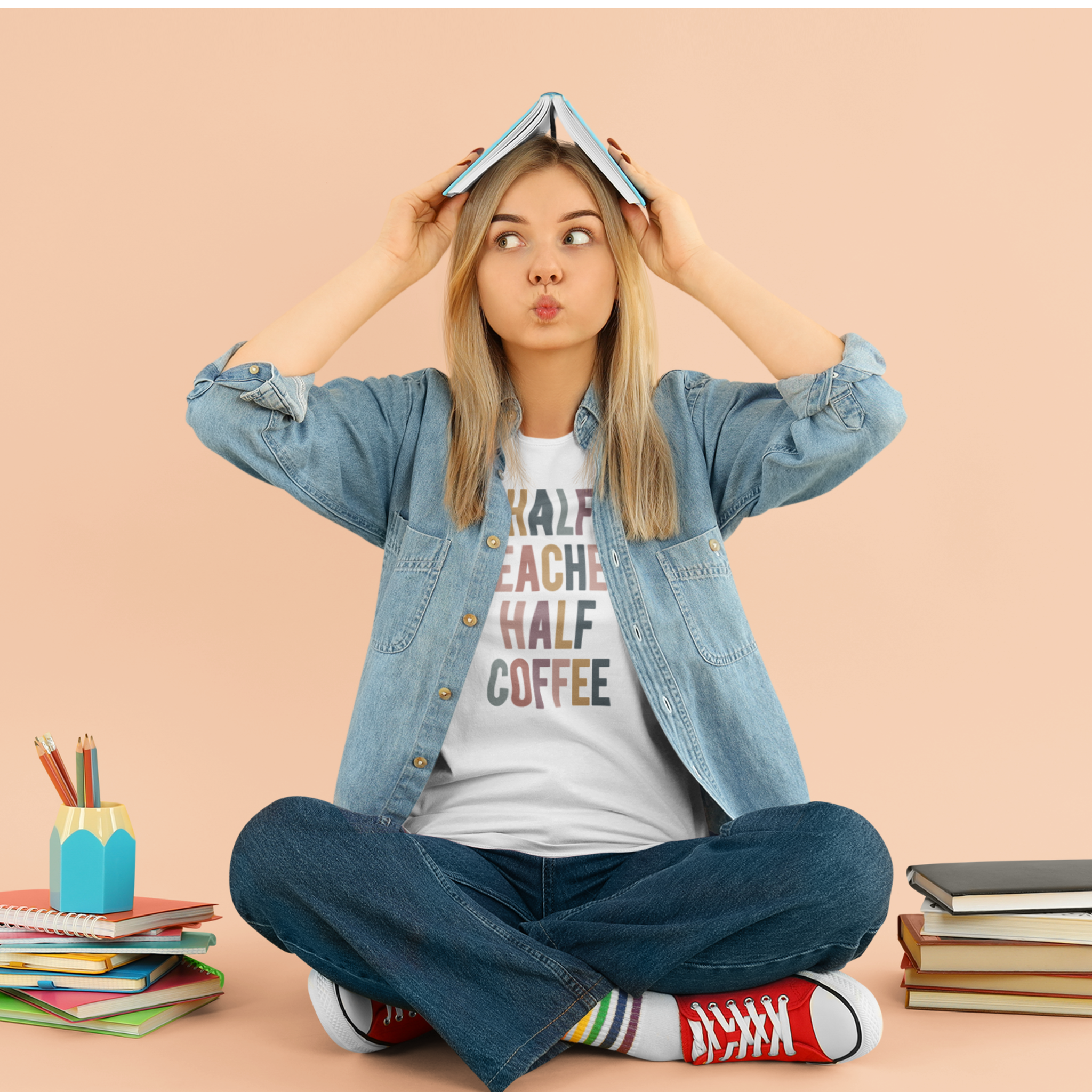 Woman sitting cross-legged on the floor, wearing a white "Half Teacher Half Coffee" t-shirt, denim shirt, and red sneakers, with books and school supplies around her.
