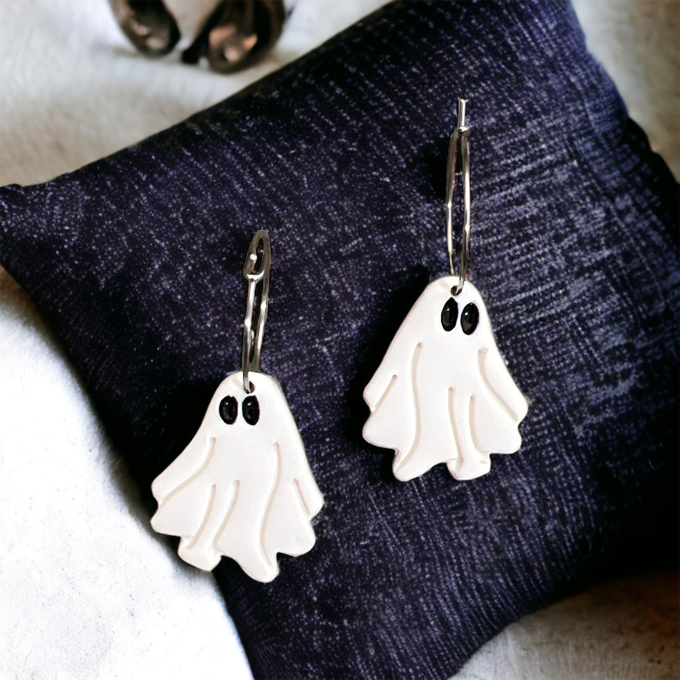 White Halloween ghost dangle earrings displayed on a soft navy pillow, highlighting their spooky charm and silver hoops. 