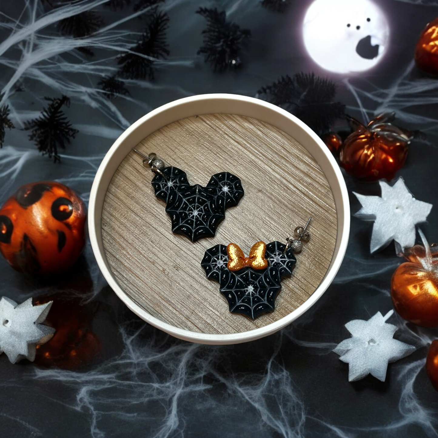 Statement spiderweb earrings with an orange glitter bow, arranged in a round display on a spooky Halloween background with festive decor. 