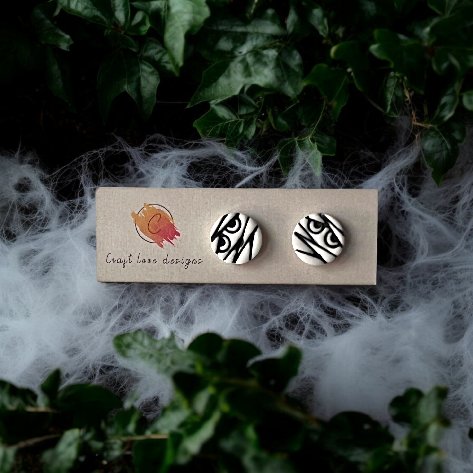 Spooky round mummy-themed stud earrings resting on a wooden tray, surrounded by cobweb decor and lush green ivy for a haunting Halloween vibe.