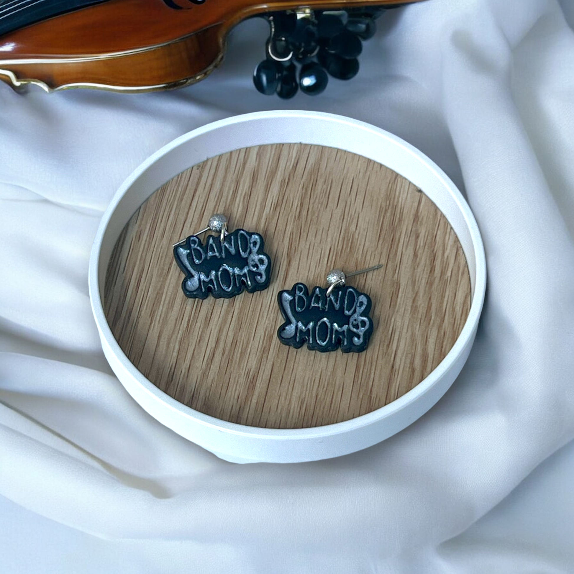 Music-themed "Band Mom" earrings with treble clef accents, resting on a wooden tray surrounded by musical instruments and white fabric. 