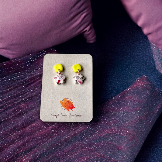 Flat lay of handmade dangle earrings featuring yellow studs and floral-patterned charms on a neutral display card with cozy fabric background.