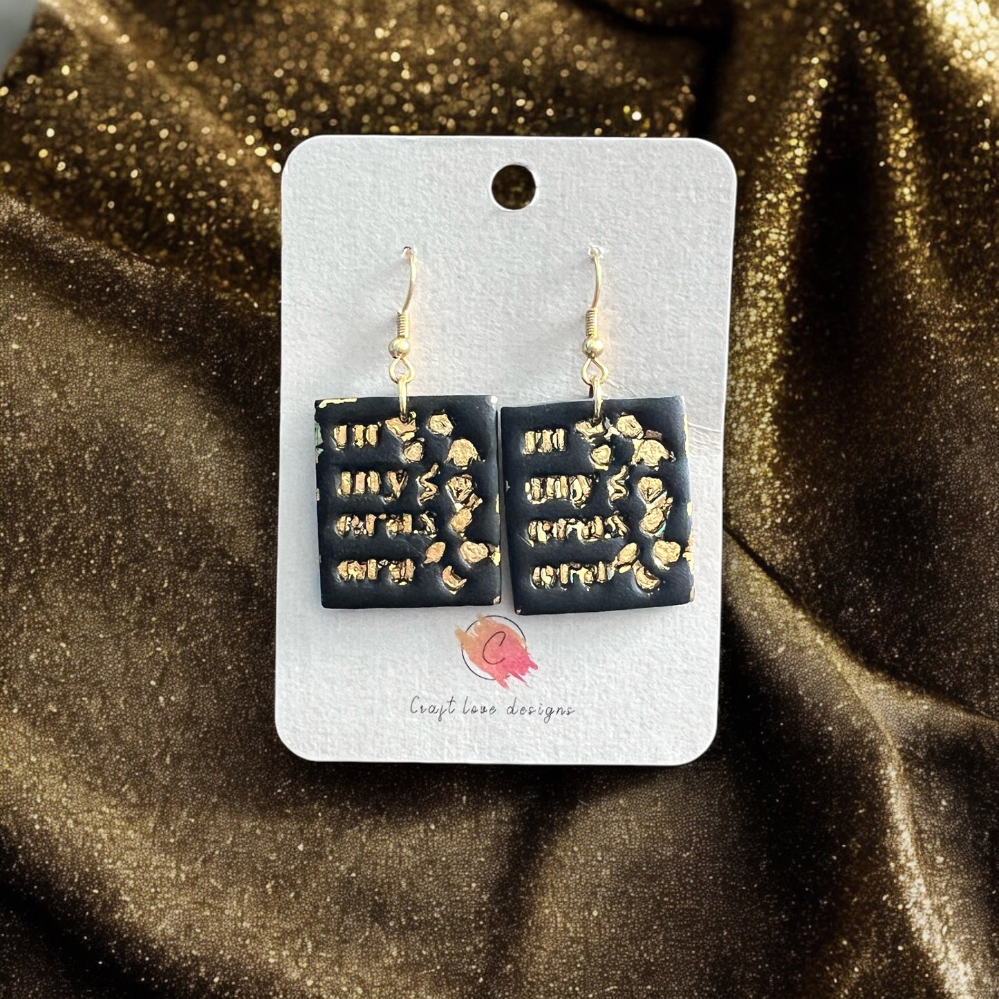 Black square dangle earrings with raised gold details displayed on a branded card against a gold fabric backdrop.