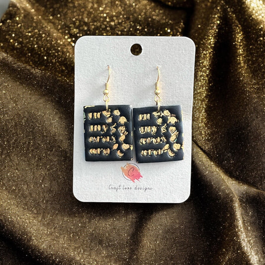 Black square dangle earrings with raised gold details displayed on a branded card against a gold fabric backdrop.