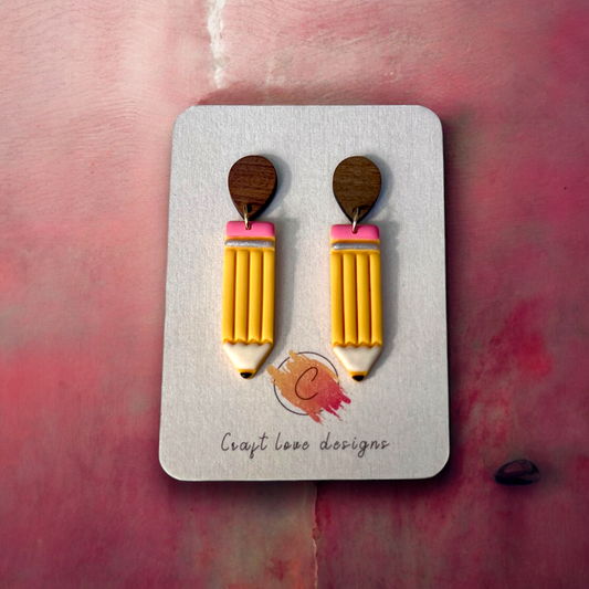 Whimsical handmade yellow pencil earrings with pink erasers, attached to wooden studs, displayed on a designer card over a soft pink background.