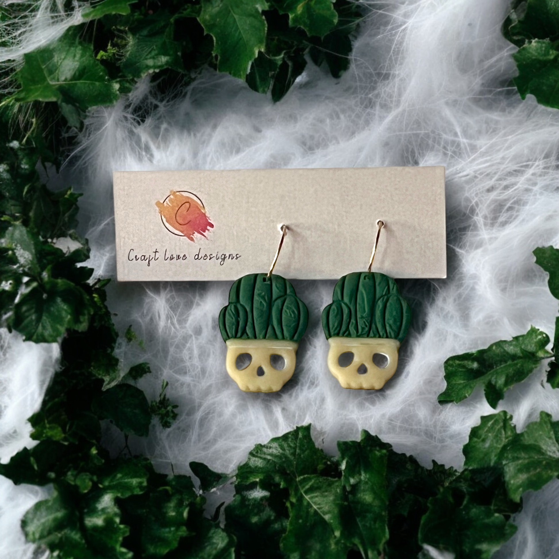 Clay dangle earrings featuring skull-shaped planters with leafy green tops, displayed on a branded card surrounded by greenery and cobwebs.