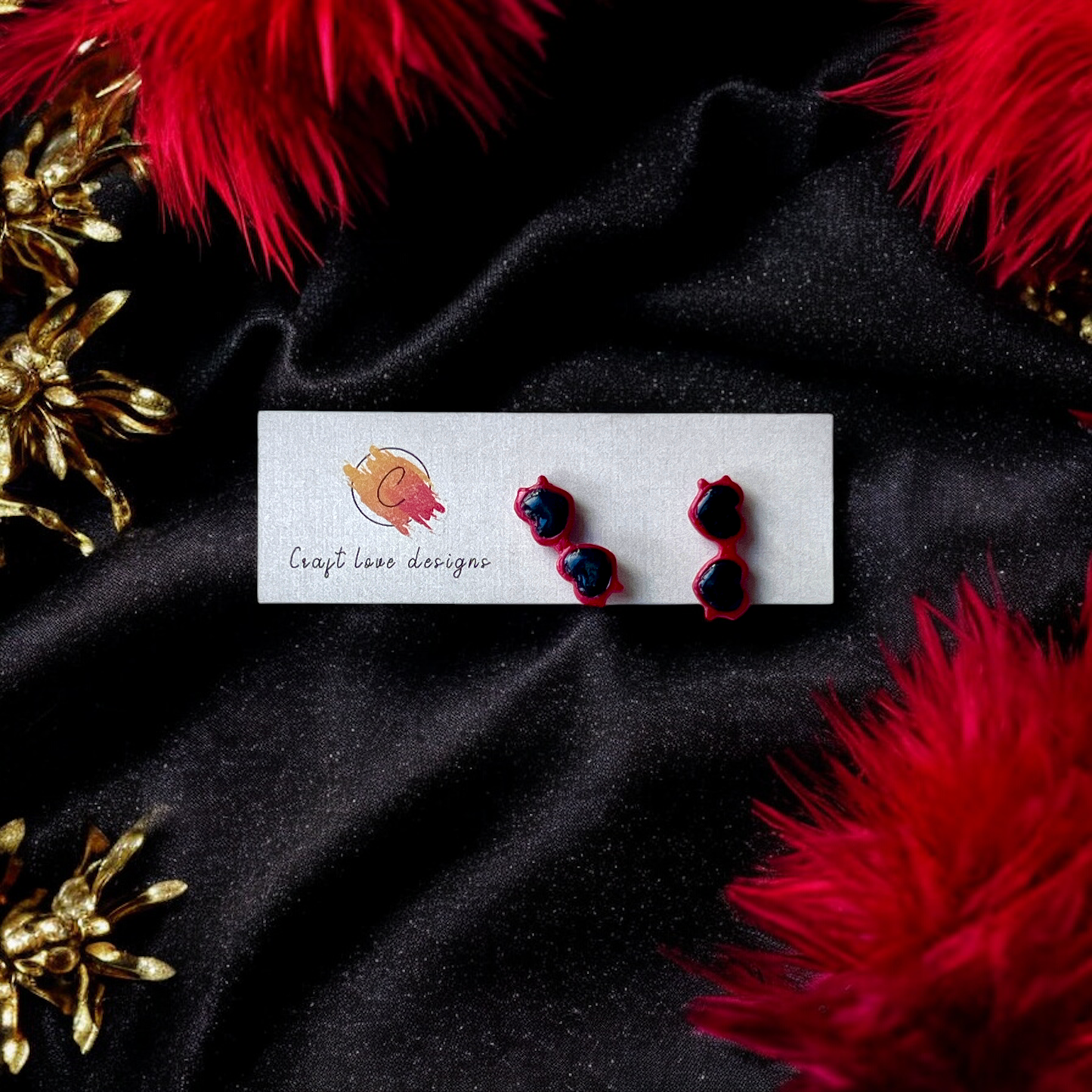Cute red heart-shaped sunglasses earrings displayed on a Craft Love Designs card with a black velvet background and red feathers.
