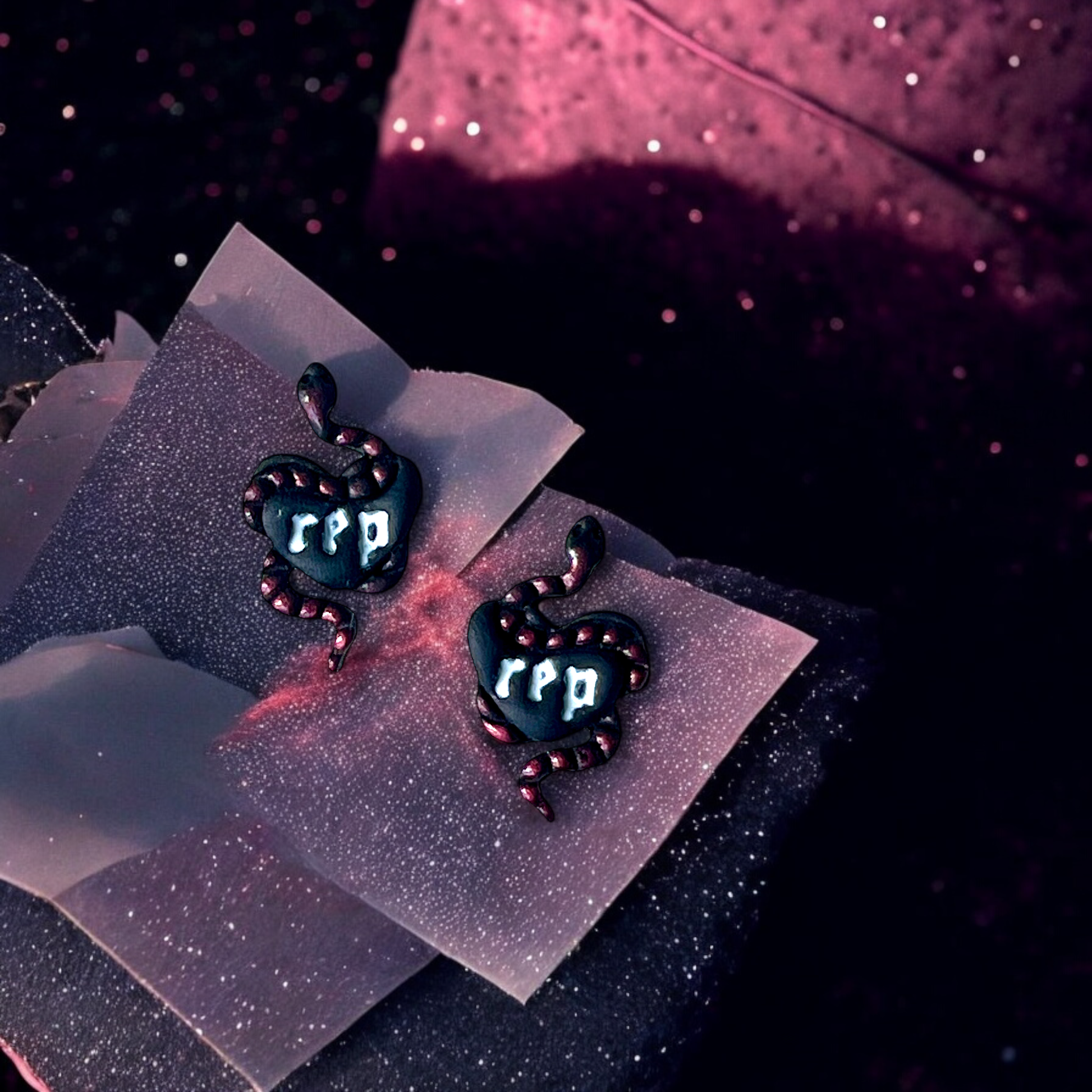 Black heart earrings wrapped in pink-detailed snakes with “rep” logo, displayed on a branded card against a cosmic pink and purple backdrop. 