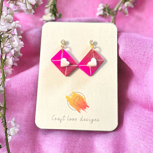 Hot pink dangle earrings with envelope design and white heart charm, creating a playful and romantic accessory. 