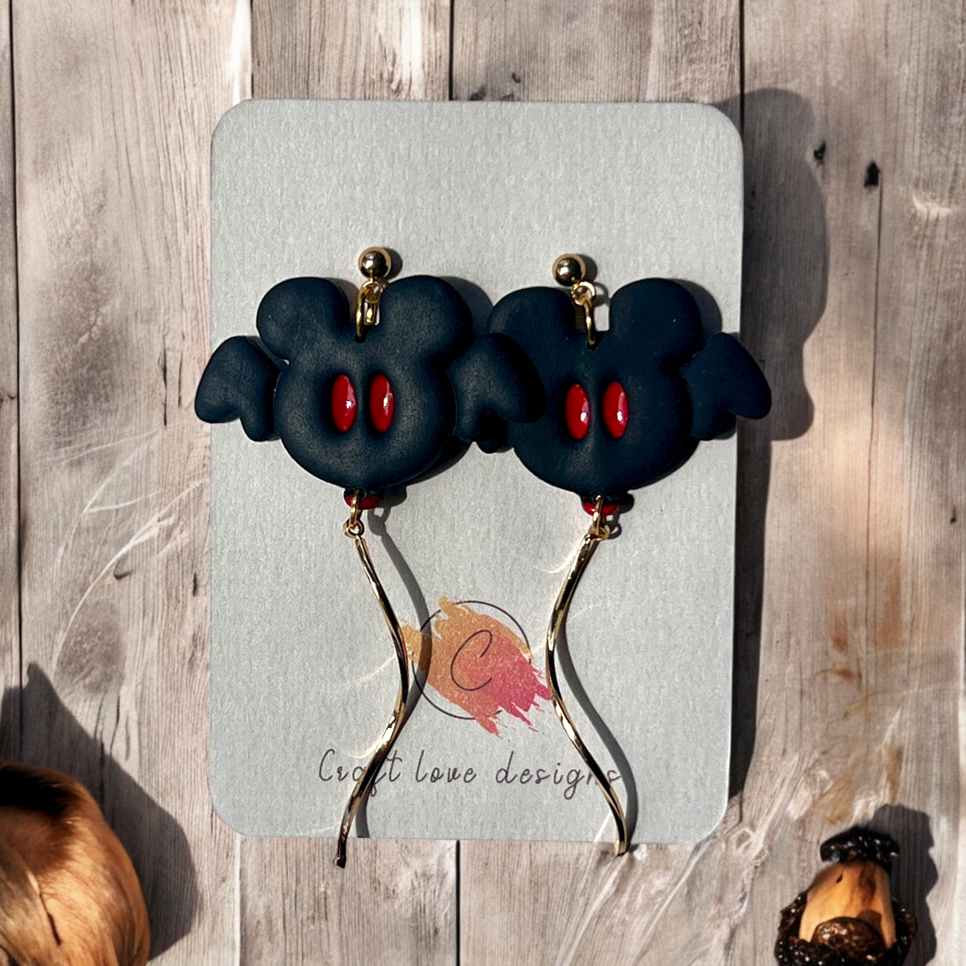 Black polymer clay bat earrings featuring red eyes and gold dangle accents, displayed on Craft Love Designs packaging.