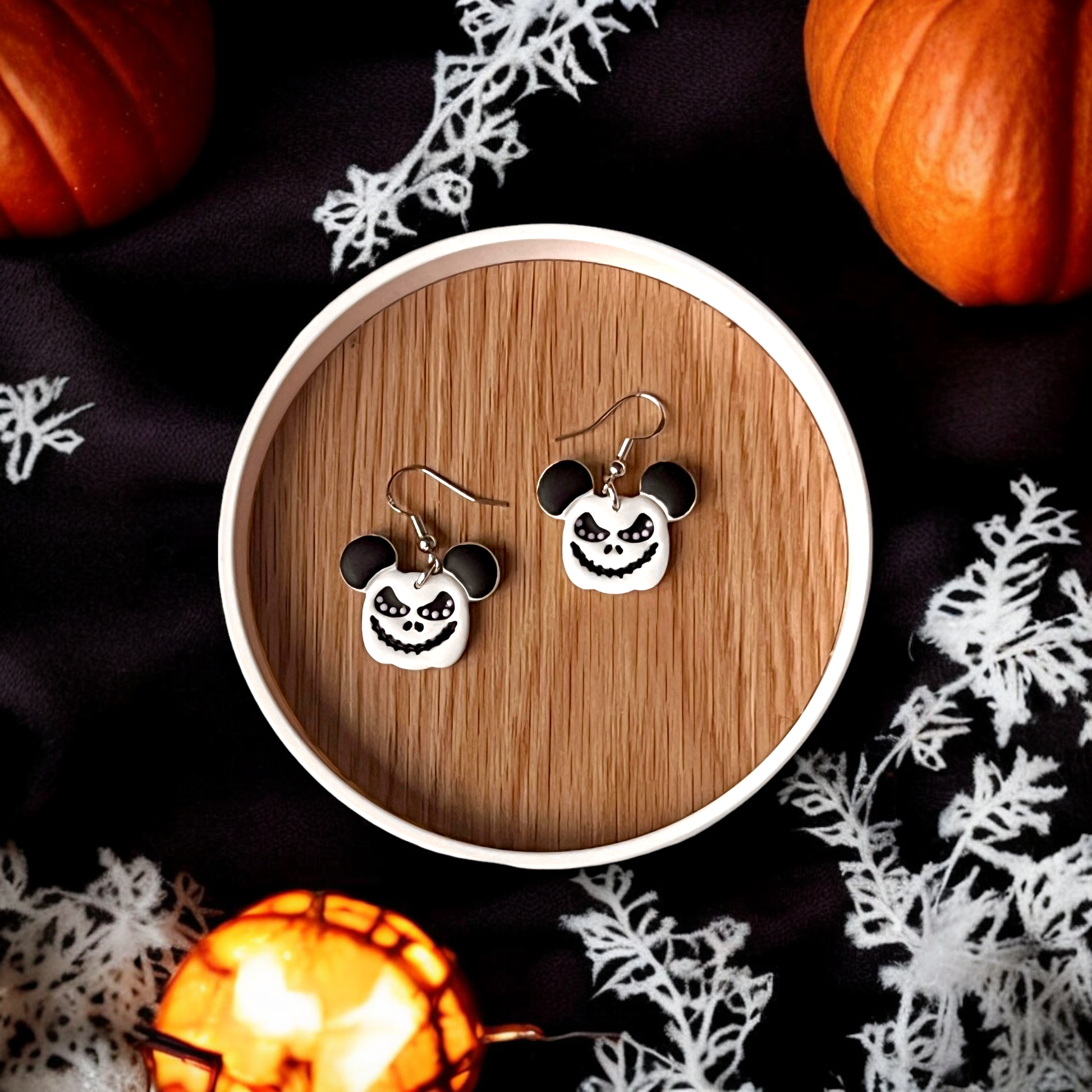 Jack Skellington-inspired earrings with Mickey Mouse ears, placed on a wooden tray with pumpkins, cobwebs, and glowing lantern decor around them.