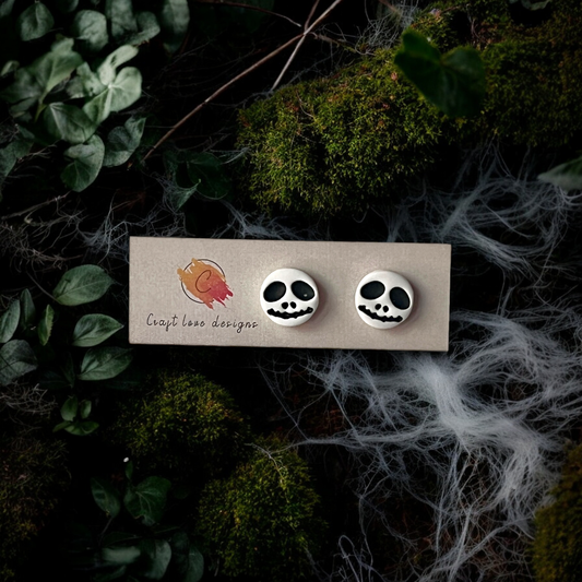 Round Jack Skellington-inspired stud earrings with spooky face design, displayed on a branded card surrounded by moss and cobwebs.