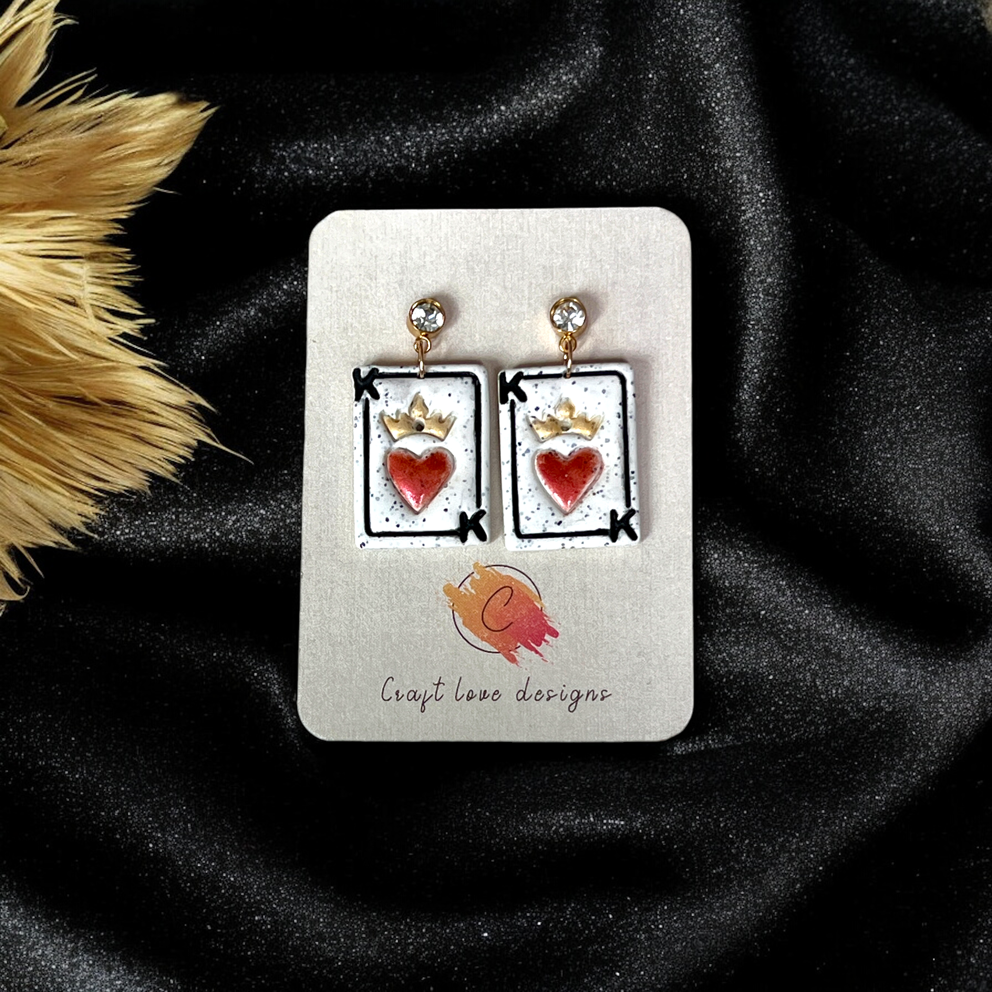 King of Hearts-inspired earrings featuring a red heart with a golden crown on a white speckled playing card design, displayed on a branded card.