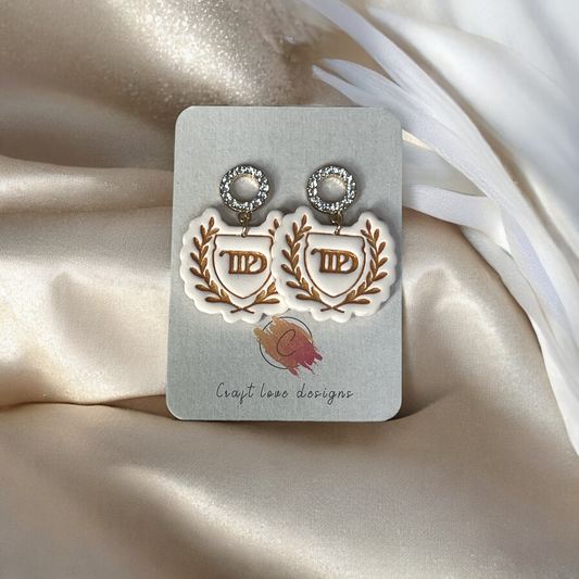 White laurel crest dangle earrings with rhinestone accents displayed on a Craft Love Designs card, set against a silky beige fabric backdrop.