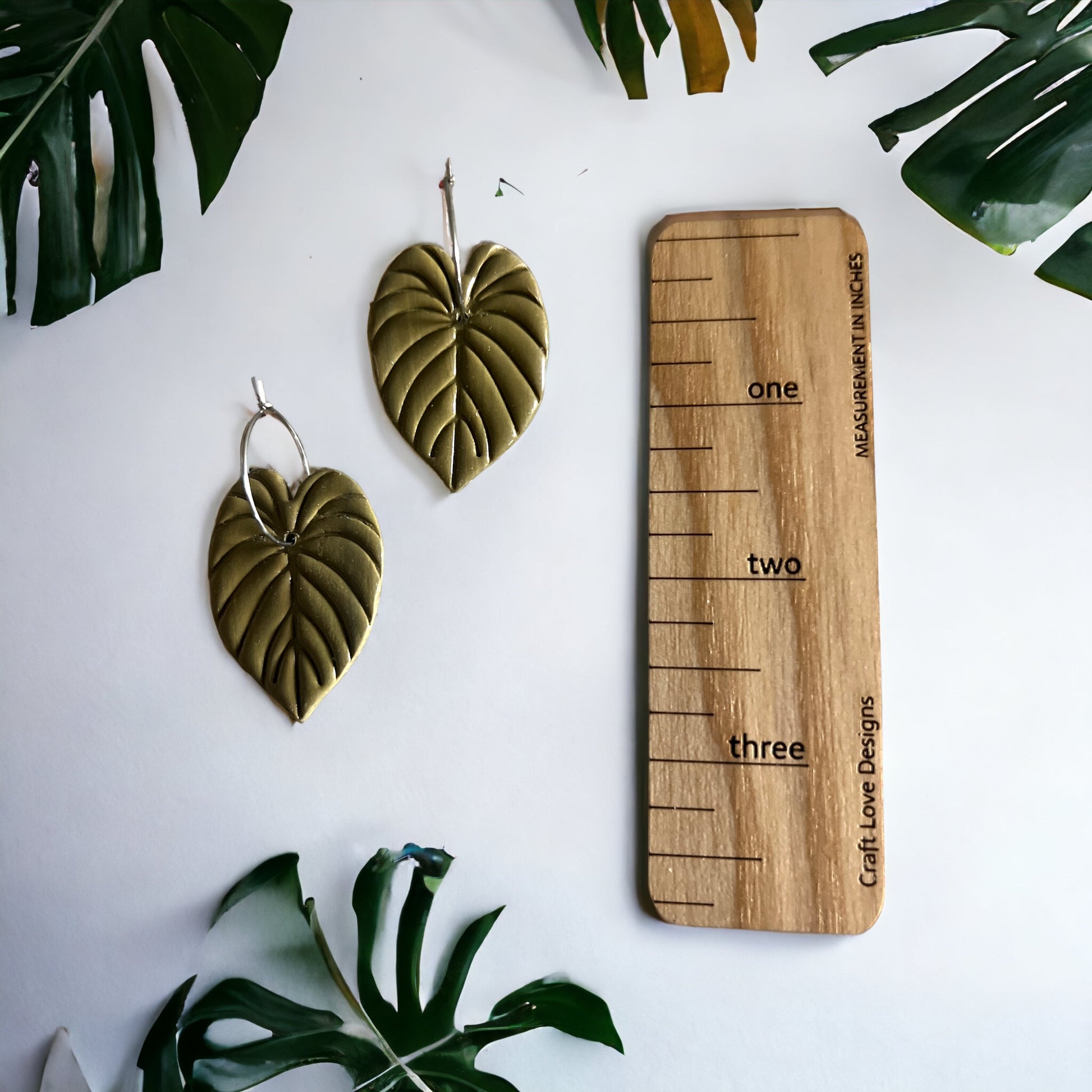 Pair of olive green philodendron leaf hoop dangle earrings displayed alongside a wooden measurement guide on a white background.