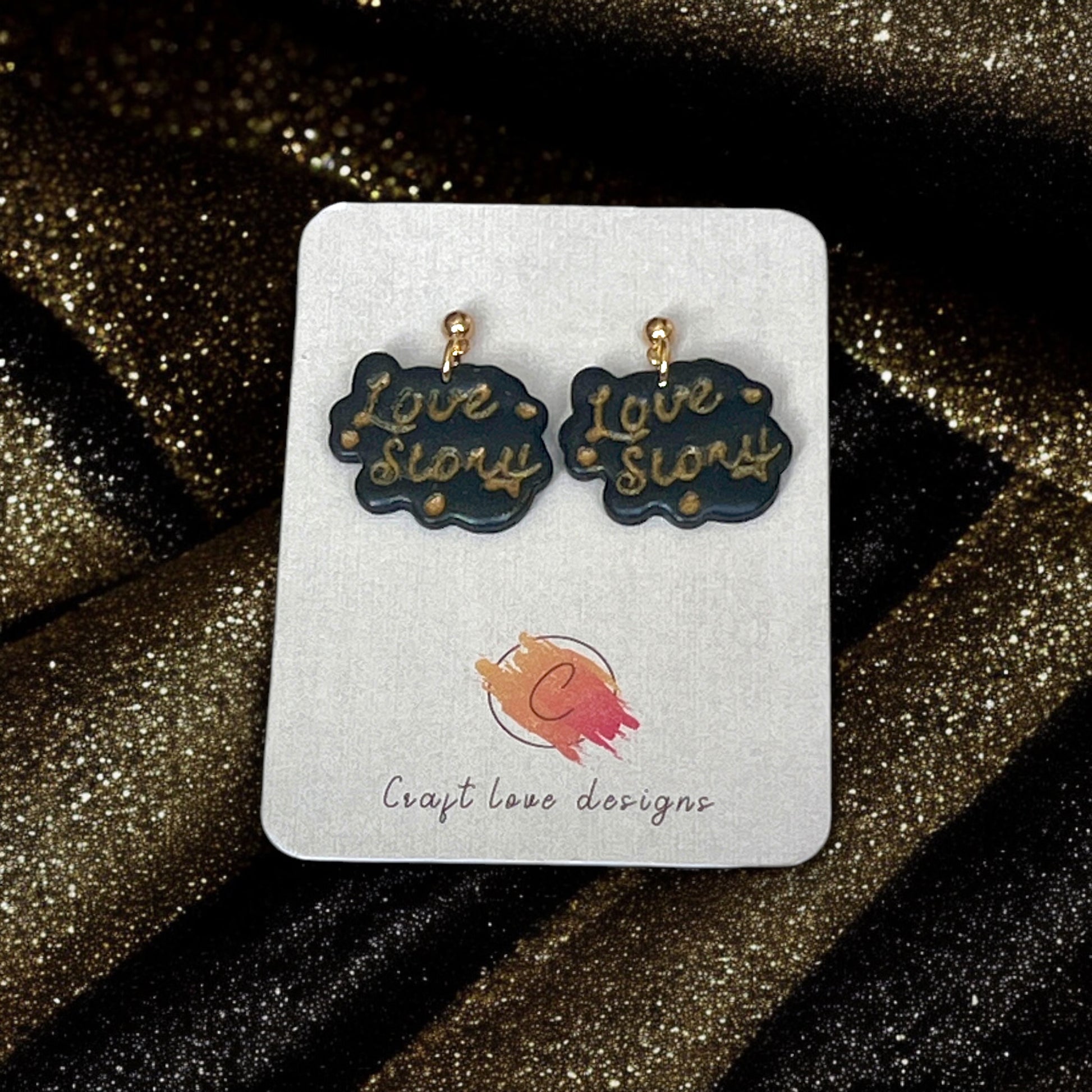 Black and gold handmade "Love Story" earrings with gold lettering, displayed on a Craft Love Designs card against sparkling black fabric.