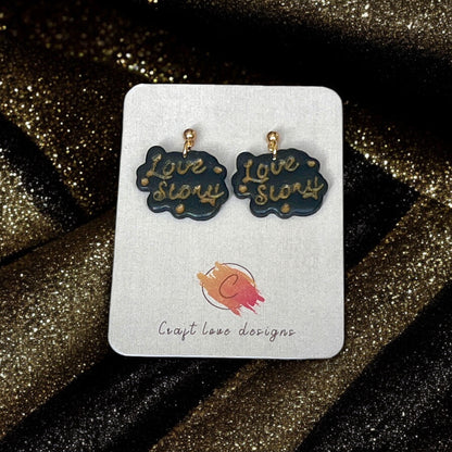 Black and gold handmade "Love Story" earrings with gold lettering, displayed on a Craft Love Designs card against sparkling black fabric.