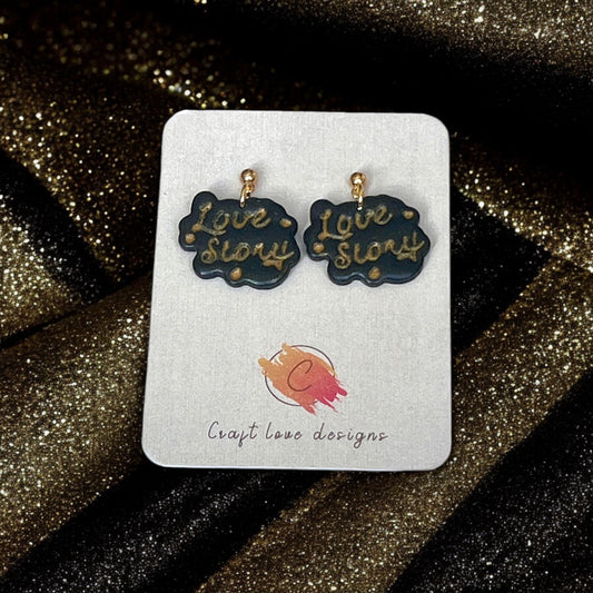 Black and gold handmade "Love Story" earrings with gold lettering, displayed on a Craft Love Designs card against sparkling black fabric.