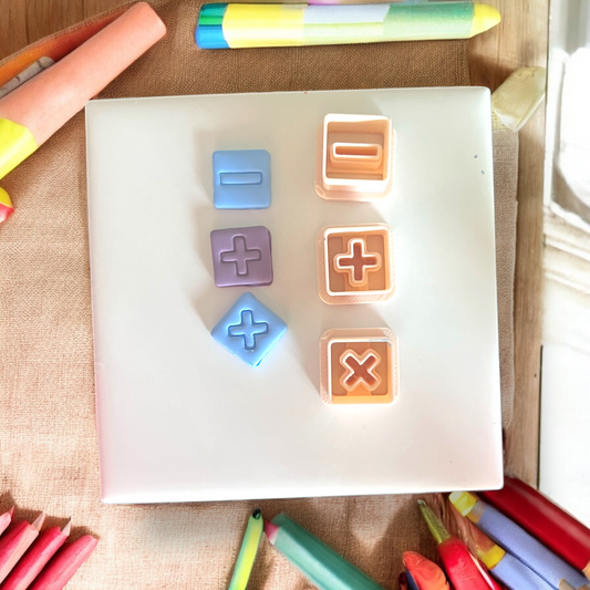 Full view of pastel orange math symbol cutters next to matching clay impressions on a white square board.