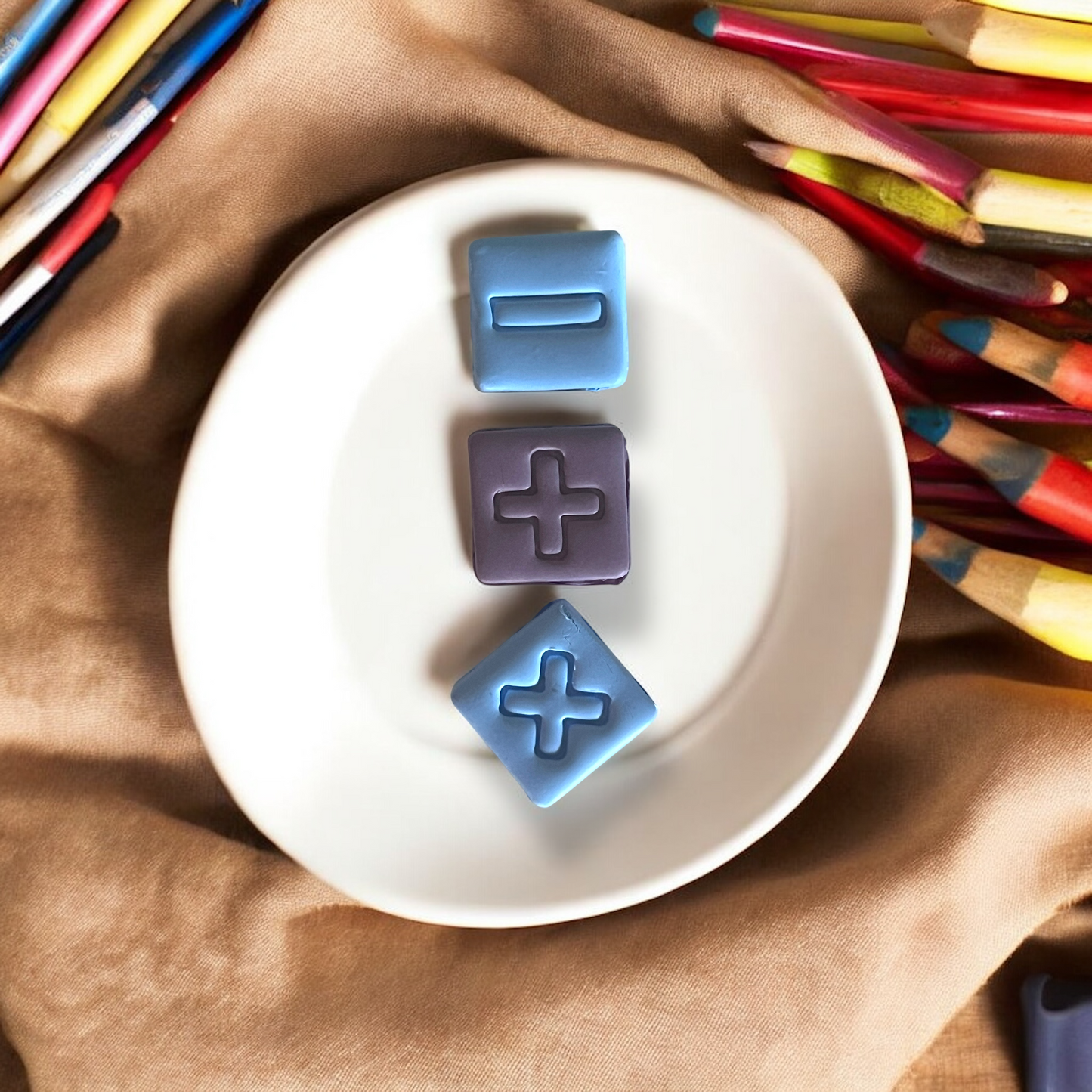 Close-up of three clay math icons on a round white dish with colored pencils and fabric background.