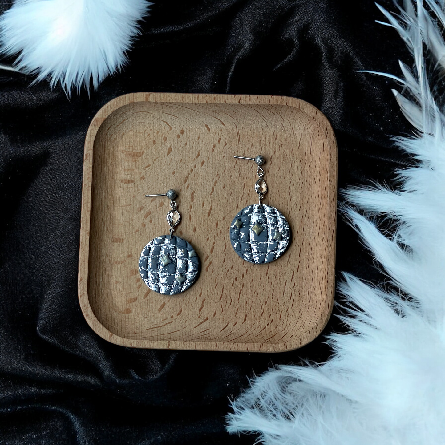 Metallic silver textured circle earrings featuring raised stars and crystal dangles, placed on a wooden tray with soft white feathers and black fabric.