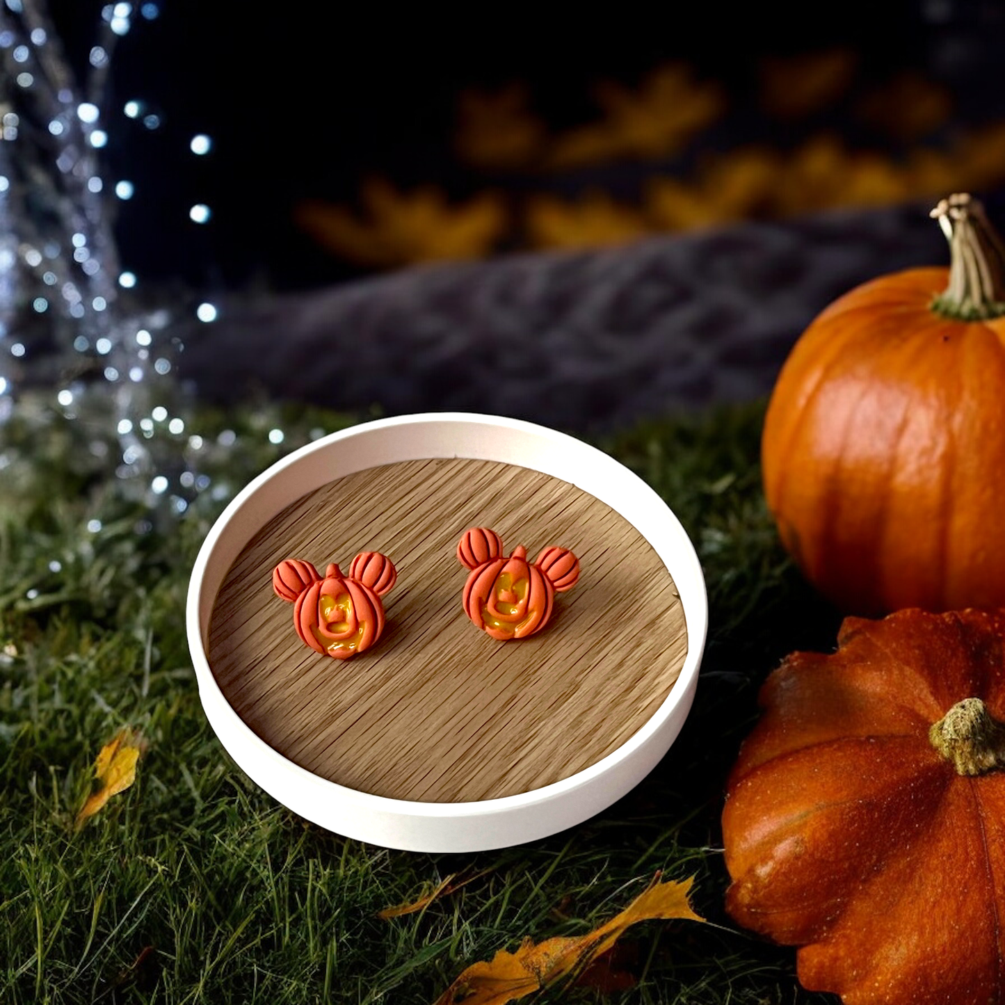 Mickey-inspired pumpkin stud earrings resting on a wooden tray, surrounded by pumpkins, grass, and warm autumn scenery with glowing lights.