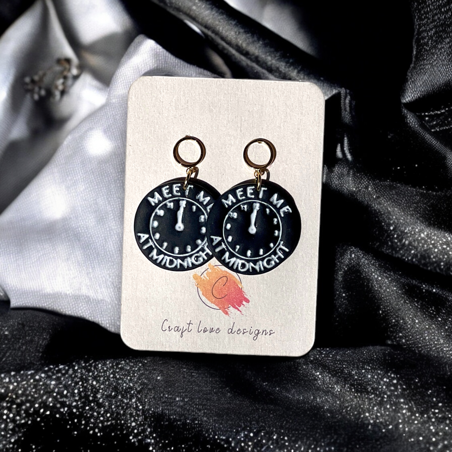 Circular midnight clock-themed drop earrings with gold rings, featuring "Meet Me At Midnight" text, displayed on a jewelry card against shiny fabric.