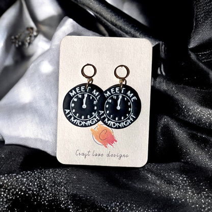 Circular midnight clock-themed drop earrings with gold rings, featuring "Meet Me At Midnight" text, displayed on a jewelry card against shiny fabric.