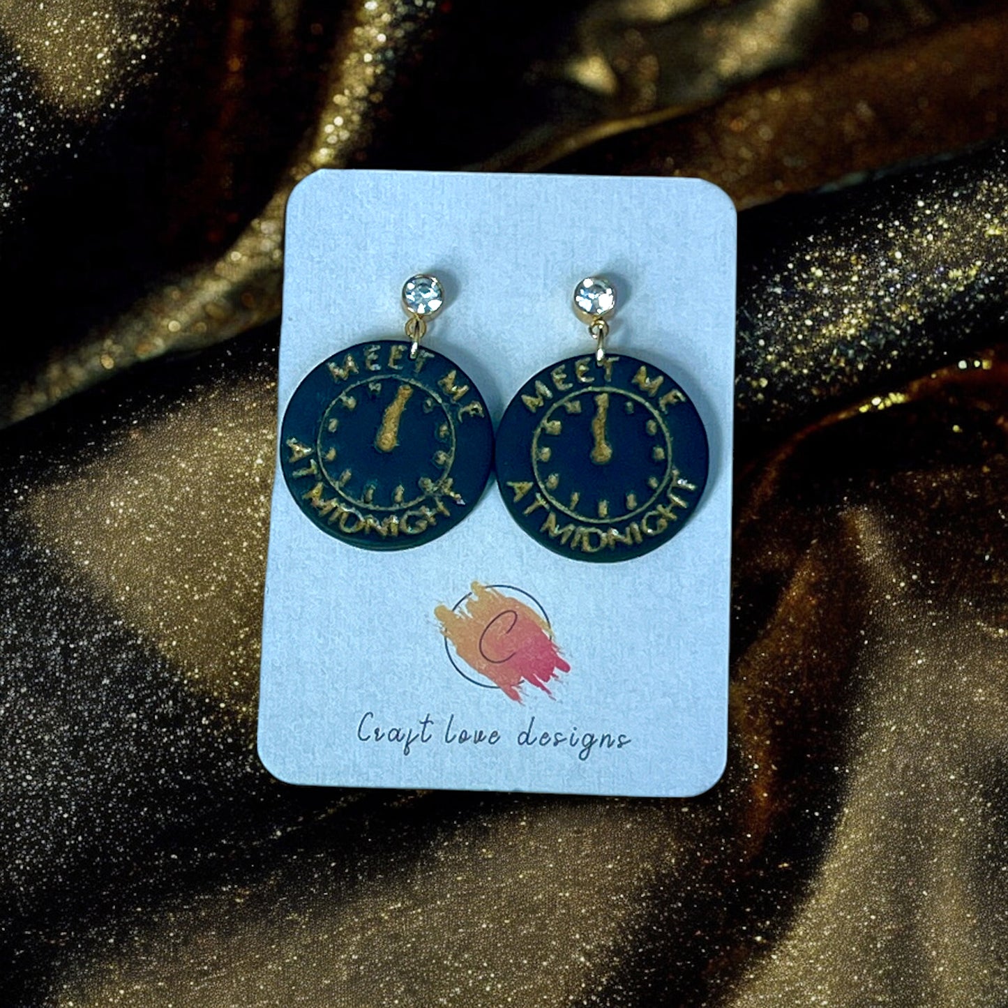 Round earrings featuring a gold midnight clock design with "Meet Me At Midnight" text, set on a jewelry card with sparkling gold fabric.