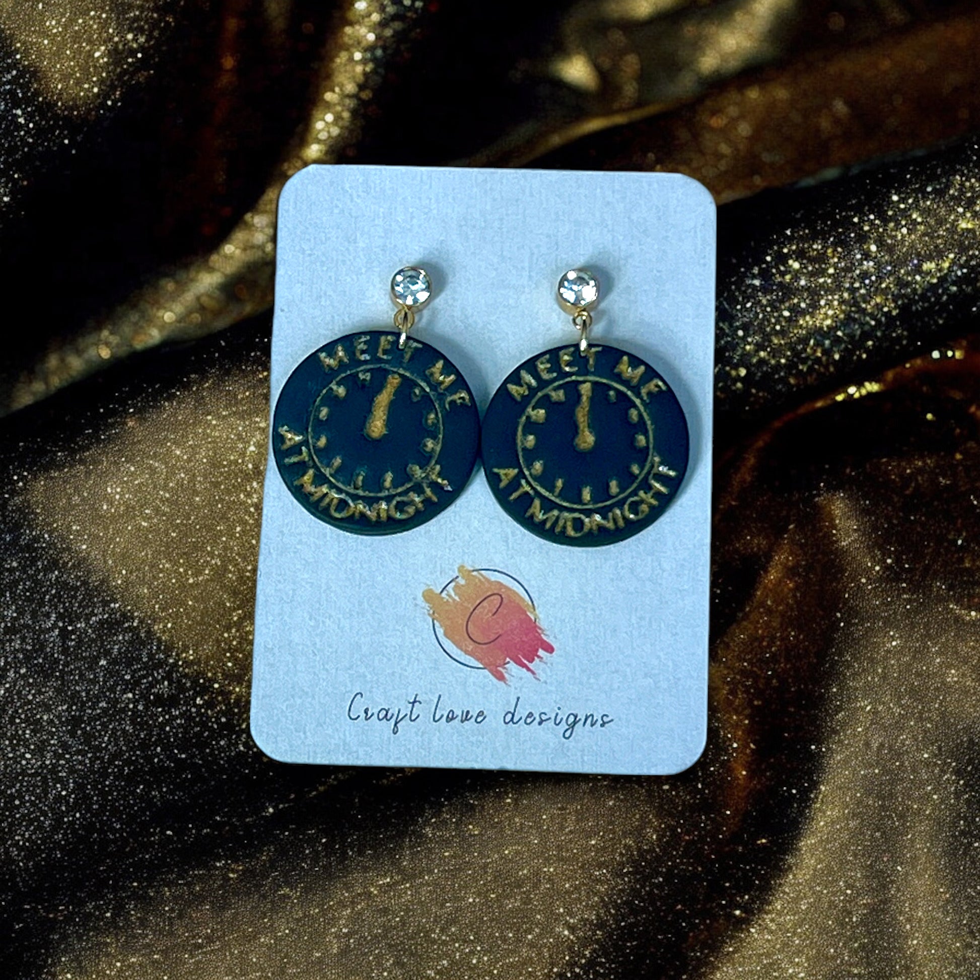 Round earrings featuring a gold midnight clock design with "Meet Me At Midnight" text, set on a jewelry card with sparkling gold fabric.