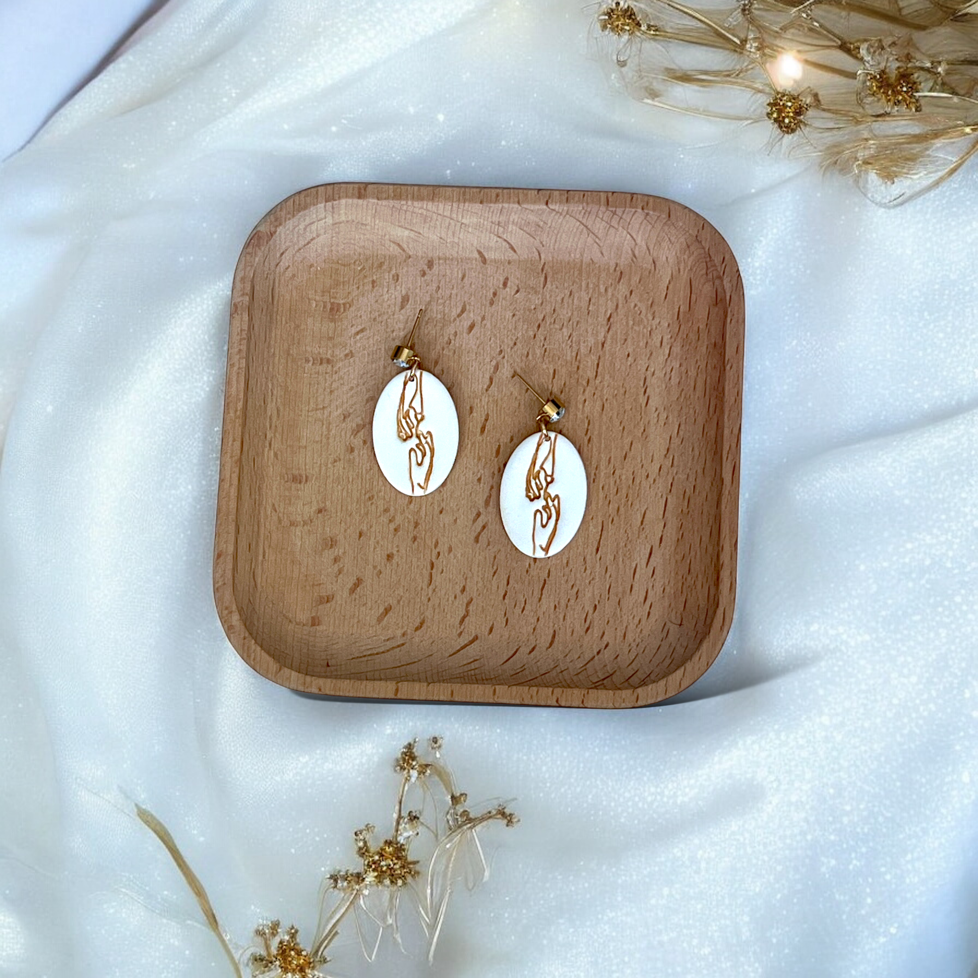 Cream oval earrings with delicate engraved gold hand line art, placed on a wooden tray surrounded by soft white fabric and golden dried florals. 