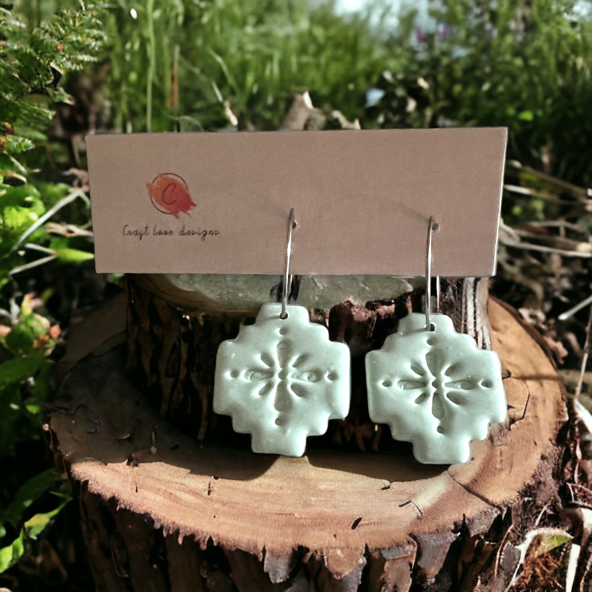 Close-up of mint green Aztec-inspired dangle earrings on hoop wires, displayed on a wooden stump amidst lush greenery.