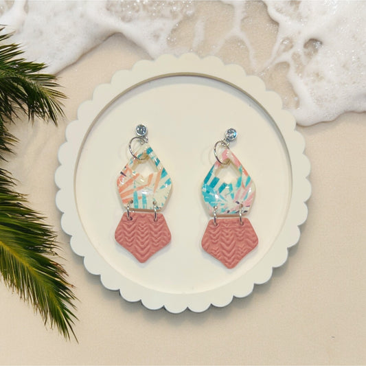 Multicolor clay dangle earrings with textured polymer clay accents on a scalloped white tray, displayed on a beach background.