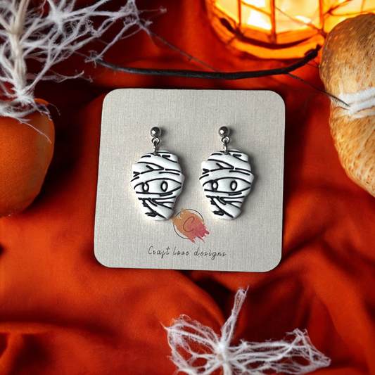 Spooky white mummy face earrings with black bandage details, displayed on a card against an orange Halloween-themed background.