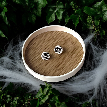 Round black-and-white mummy face stud earrings with spooky bandage design, displayed on a branded card surrounded by cobwebs and ivy.