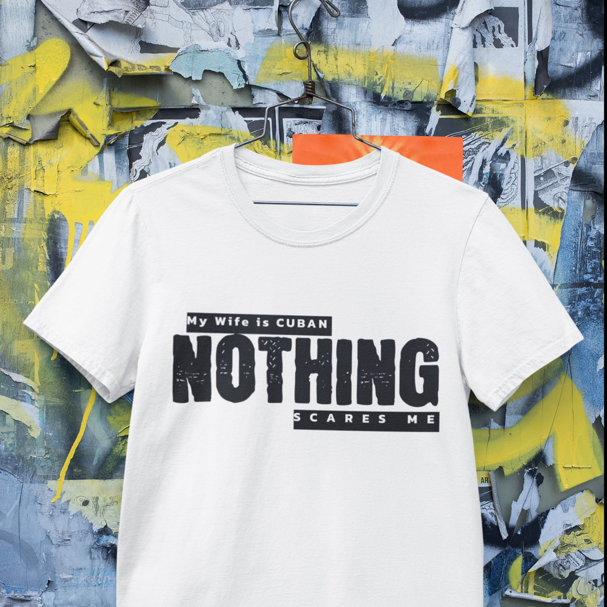 "My Wife is Cuban, Nothing Scares Me" t-shirt hanging on a hanger, styled against a colorful wall setting.