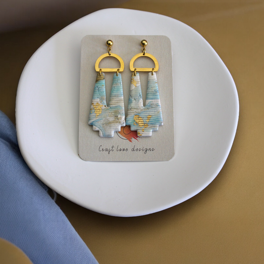 Ocean-inspired drop earrings with marbled blues and gold accents, resting on a neutral ceramic dish. Simple, modern, and beachy. 