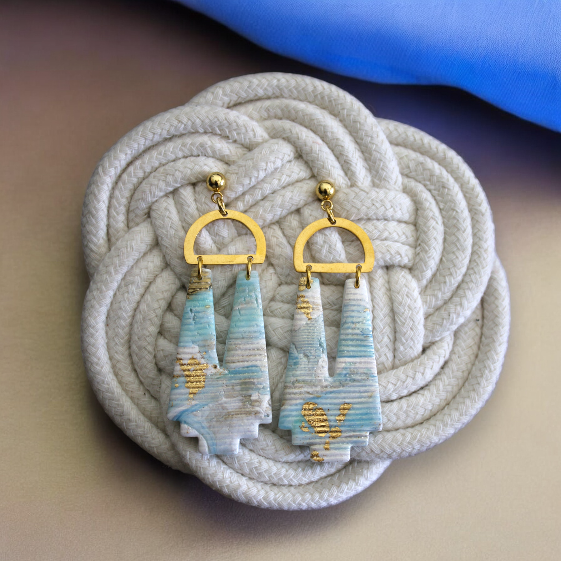 Handmade marbled earrings in soft ocean-inspired hues with gold accents, displayed on a wooden stand with warm lighting. 