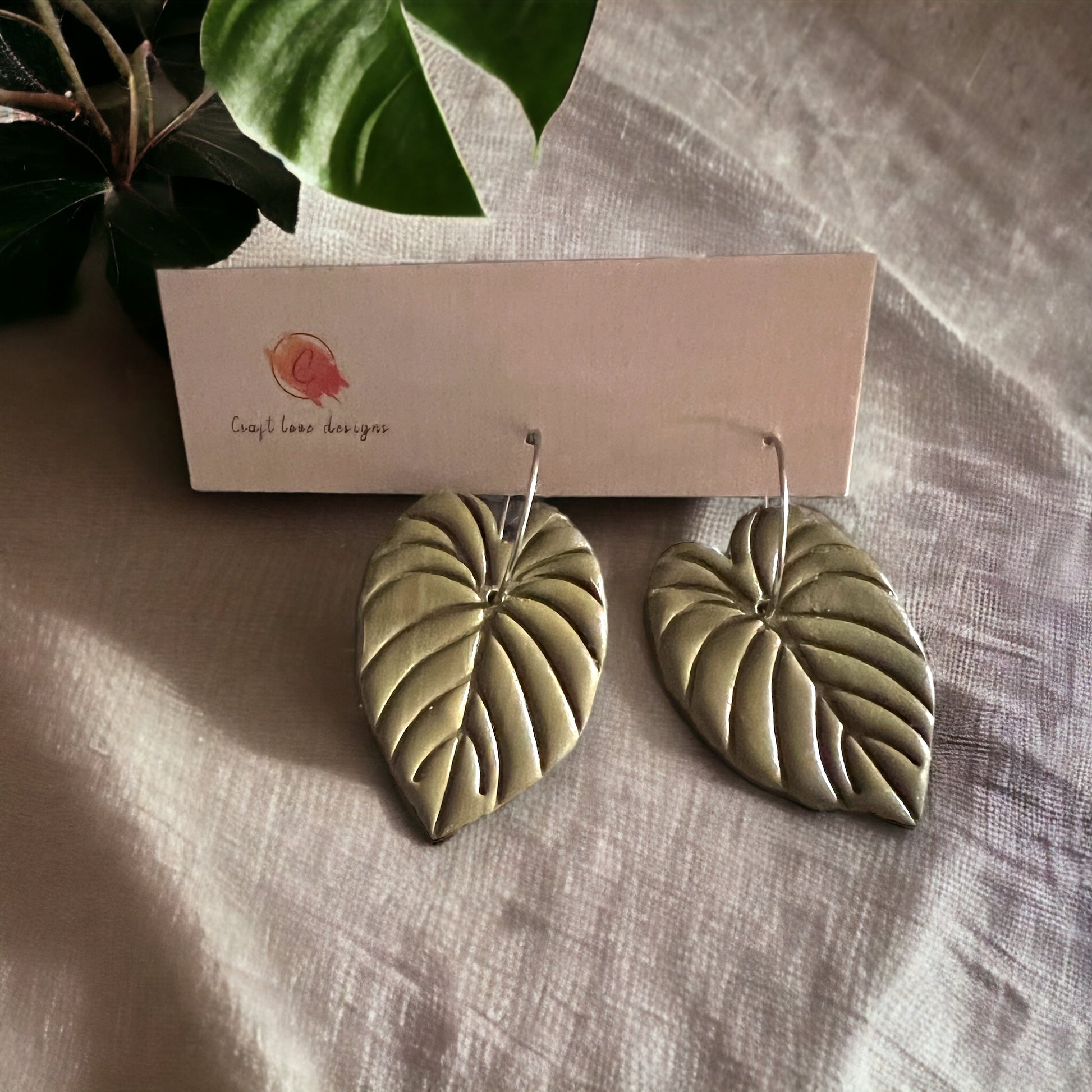 Close-up view of olive green philodendron leaf hoop dangle earrings by Craft Love Designs on a neutral surface.