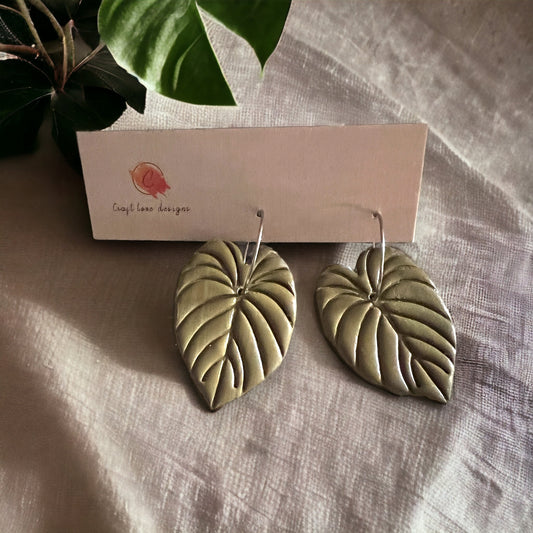 Close-up view of olive green philodendron leaf hoop dangle earrings by Craft Love Designs on a neutral surface.