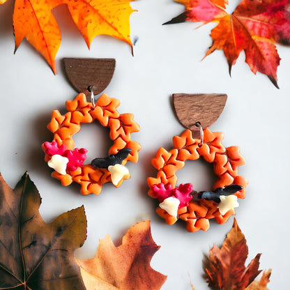 A close-up of orange wreath dangle earrings, featuring autumn leaves, berries, and bats, with warm seasonal tones in the background.